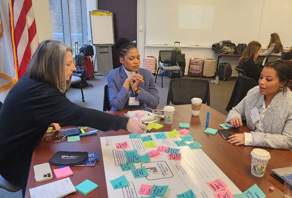 Three women sit discussing ideas at a conference table covered with stick notes